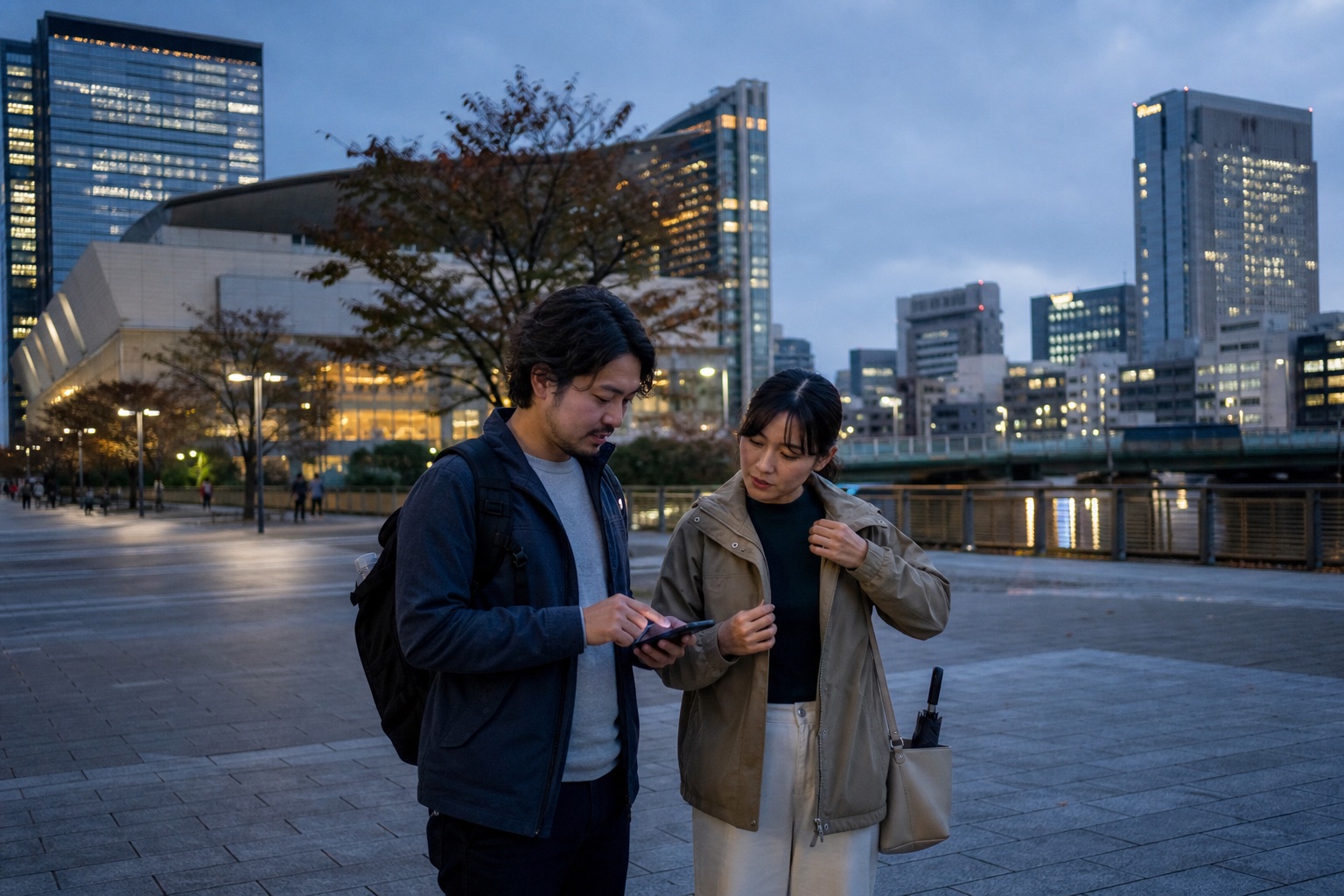 Travelers in light jackets checking their route beside a Tokyo waterfront at blue hour