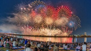 A large fireworks display over the Tama River with spectators on the Chofu riverbank