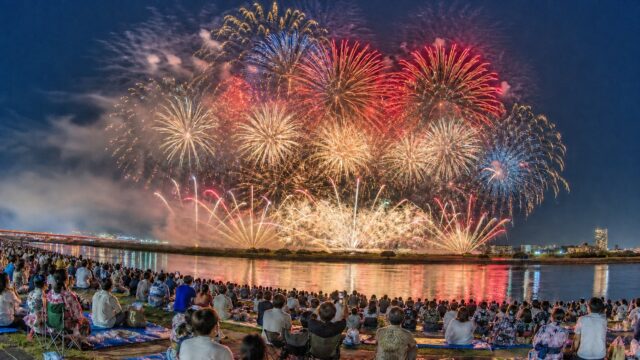 A large fireworks display over the Tama River with spectators on the Chofu riverbank