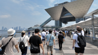 Visitors carrying water walk toward a waterfront exhibition venue on a hot summer morning