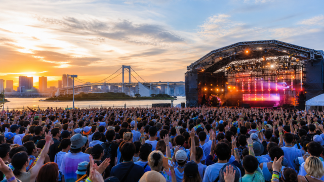 An outdoor electronic music festival crowd on the Odaiba waterfront at sunset