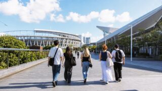 International music fans walking through a sunny coastal event district near a stadium and exhibition halls