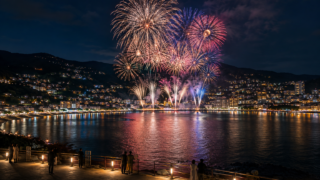 AI-generated editorial illustration of fireworks over a mountain-framed coastal bay